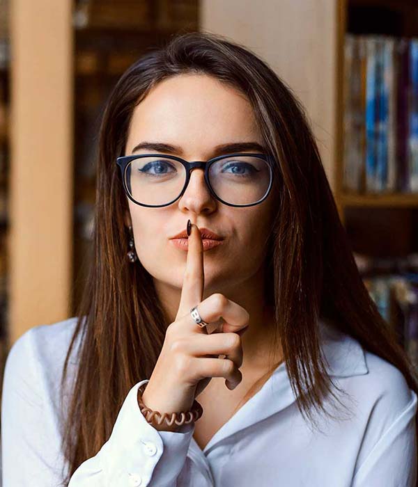 Female San Jacinto College Student holds a finger to her lips in a "shh" gesture, standing in front of library bookshelves.